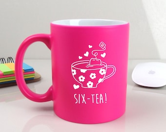 May include: A bright pink ceramic mug with a white outline of a teacup with a teabag and the text "SIX-TEA!"