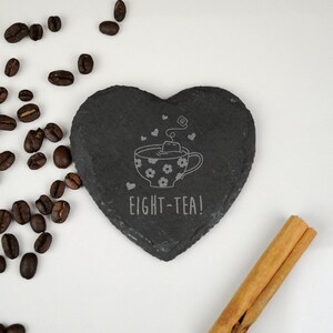May include: A black heart-shaped slate coaster with a white design of a teacup and the text "EIGHT-TEA!"