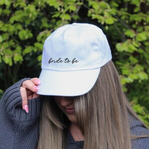 May include: White baseball cap with the text "bride to be" embroidered in black on the front.