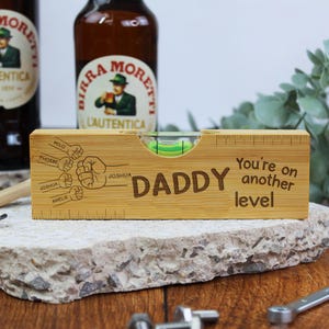 Puede incluir: Un nivel de madera con la palabra "DADDY" grabada, junto con la frase "You're on another level". El nivel tiene una burbuja y marcas de regla. Los nombres están grabados cerca de dibujos de puños.