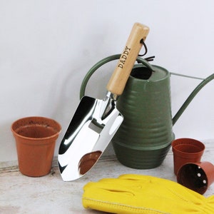 Pode incluir: Uma pá de jardim de prata com um cabo de madeira gravado com a palavra "DADDY". A pá está ao lado de um regador verde e um vaso de flores castanho.