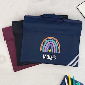 May include: Three school book bags in navy, black, and burgundy. The navy bag has a rainbow design and the name "Maisie". The bags have a handle and a flap closure. The bags are suitable for school.