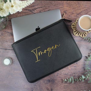 May include: Black laptop sleeve with a gold zip and the name "Imogen" in gold script. A silver laptop is partially visible inside the sleeve. The sleeve is on a wooden surface, with a cup of coffee and flowers in the background.