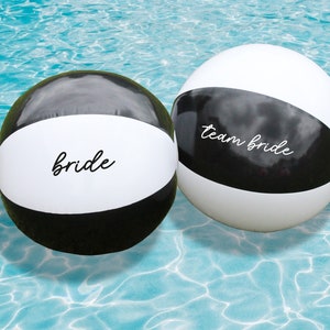 May include: Two black and white beach balls floating in a blue pool. One ball says "bride" and the other says "team bride".