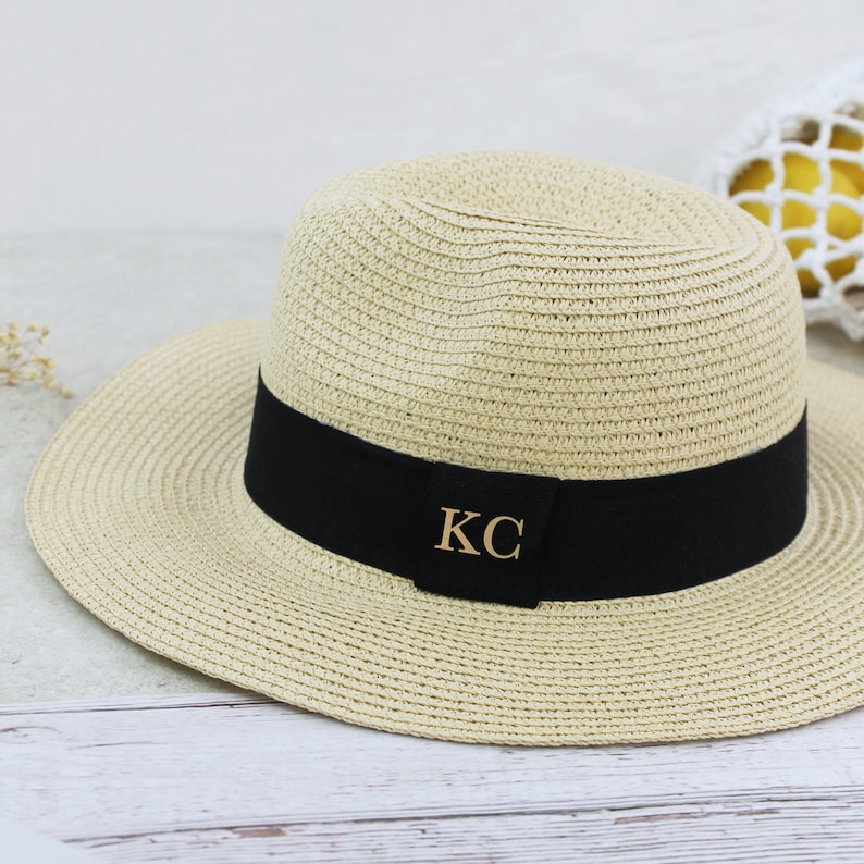 Peut inclure: Un chapeau de paille beige avec un ruban noir et les initiales dor&eacute;es "KC" sur le devant.