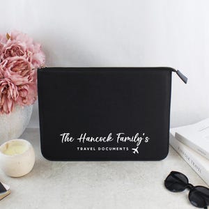May include: Black travel document holder with a zip closure. The text "The Hancock Family's TRAVEL DOCUMENTS" is printed in white, along with a small airplane graphic. A bouquet of pink flowers and a candle are in the background.