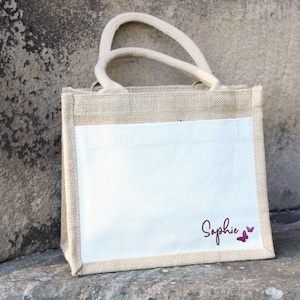 May include: A natural jute tote bag with a white cotton panel. The panel has the name "Sophie" written in cursive with two small pink butterflies.