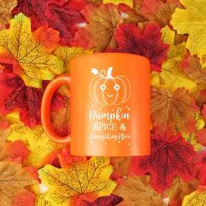 May include: An orange ceramic mug with a white pumpkin graphic and the text "Pumpkin Spice & Everything Nice." The mug is surrounded by colorful autumn leaves, creating a seasonal theme.