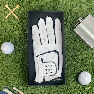 K&ouml;nnte beinhalten: Ein Golf-Geschenkset auf gr&uuml;nem Gras. Das Set enth&auml;lt einen wei&szlig;en Golfhandschuh in einer schwarzen Box, eine silberne Flasche, zwei Golfb&auml;lle und zwei Holz-Tees. Der Golfhandschuh hat einen schwarzen Akzent und die Initialen "TF" mit gekreuzten Golfschl&auml;gern.
