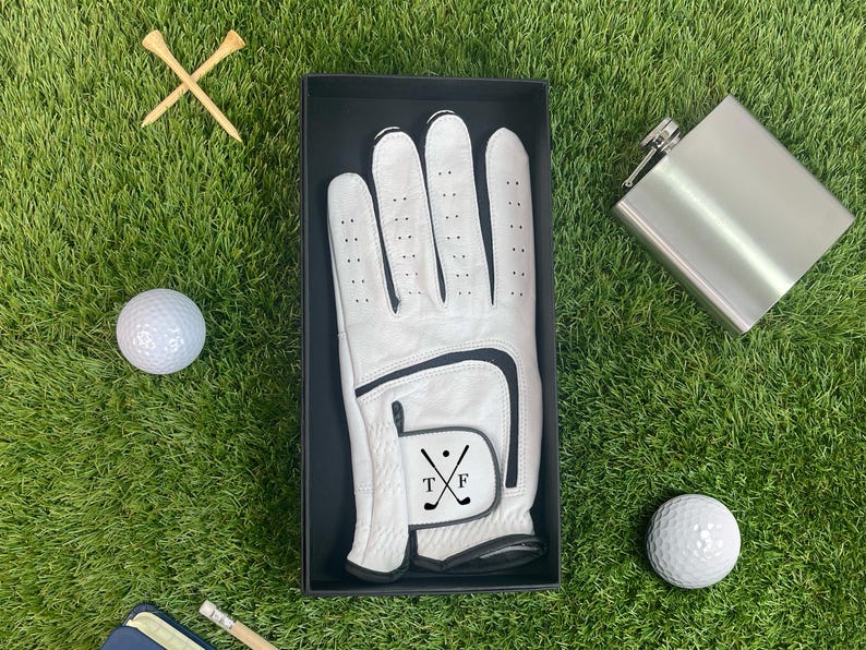 K&ouml;nnte beinhalten: Ein Golf-Geschenkset auf gr&uuml;nem Gras. Das Set enth&auml;lt einen wei&szlig;en Golfhandschuh in einer schwarzen Box, eine silberne Flasche, zwei Golfb&auml;lle und zwei Holz-Tees. Der Golfhandschuh hat einen schwarzen Akzent und die Initialen "TF" mit gekreuzten Golfschl&auml;gern.