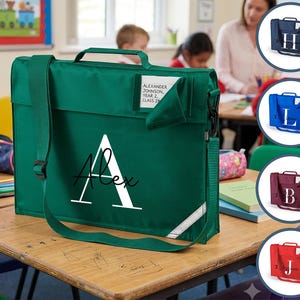 May include: A rectangular green school book bag with a top handle and an adjustable shoulder strap. The front features a large white letter A with the name Alex printed in black script. A clear name tag pocket contains a card with printed text.