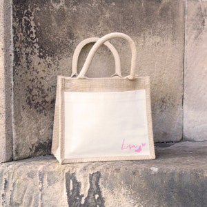 May include: A natural jute tote bag with a white fabric insert. The bag has a pink butterfly design and the name "Lisa" written on the front.