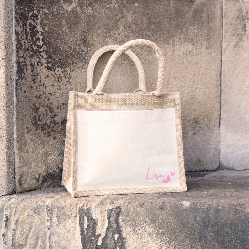 May include: A natural jute tote bag with a white fabric insert. The bag has a pink butterfly design and the name "Lisa" written on the front.