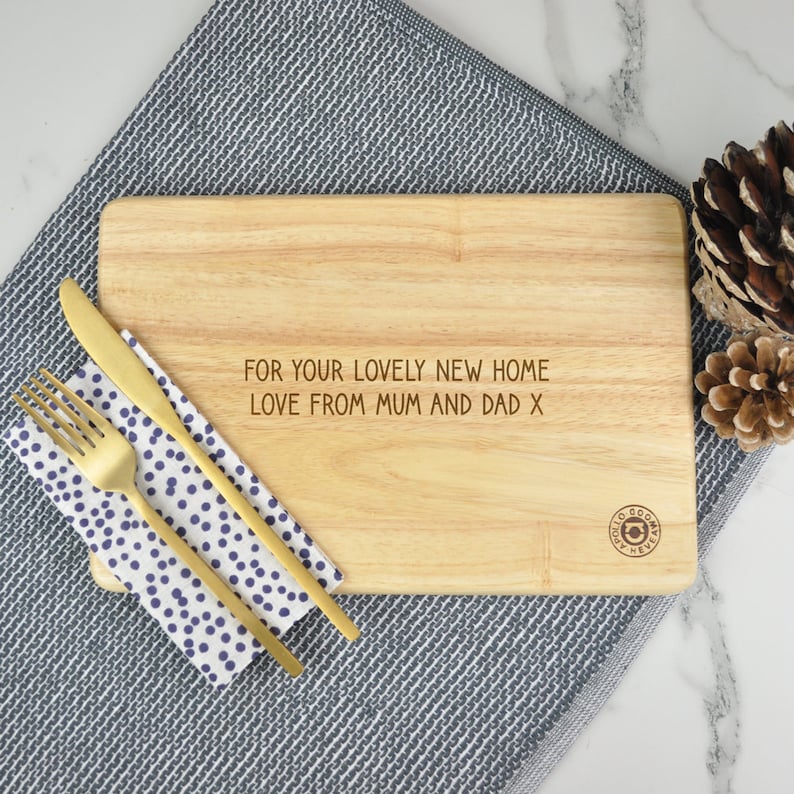 May include: A wooden cutting board with the text "For your lovely new home Love from Mum and Dad x" engraved on it. The board is sitting on a gray and white patterned surface.