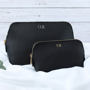 May include: Two black cosmetic bags with gold zippers and personalized initials. The larger bag has "O.E" and the smaller bag has "V.B" in gold lettering. The bags are displayed on a white satin surface.