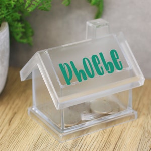 Pode incluir: Um cofrinho de plástico transparente em forma de casa com o nome "Phoebe" escrito no telhado em letras verde-azuladas. O cofrinho está sobre uma superfície de madeira e tem moedas dentro.