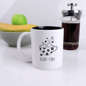 May include: White ceramic mug with a black interior and a black and white illustration of a teacup with a tea bag and the text "EIGHT-TEA!"
