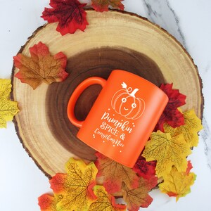 May include: An orange ceramic mug with a handle, featuring a smiling pumpkin graphic and the text "Pumpkin Spice & Everything Nice." The mug is surrounded by colorful autumn leaves on a wooden slice, creating a fall theme.