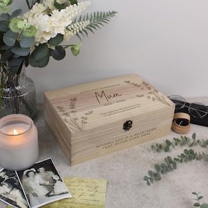 May include: A wooden memory box with the text "CHERISHED MEMORIES OF Mum 1955-2025" and a decorative lavender design. The box has a metal clasp and is surrounded by a lit candle, flowers, and old photographs.
