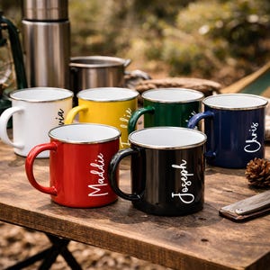May include: A selection of enamel camping mugs in different colours: white, yellow, green, blue, red, and black. Each mug has a silver rim and a black handle. Names are printed on the mugs in a white script font. The mugs are on a wooden surface.