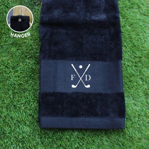 Puede incluir: Toalla de golf negra con un diseño bordado blanco con palos de golf cruzados, una pelota de golf y las iniciales "F D". La toalla tiene un lazo para colgar. Se muestra sobre una superficie verde.