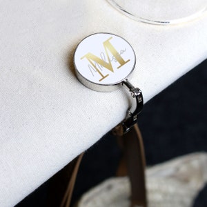 May include: A silver and white personalized bag hook with a gold letter "M" and the name "Melissa" written in cursive.