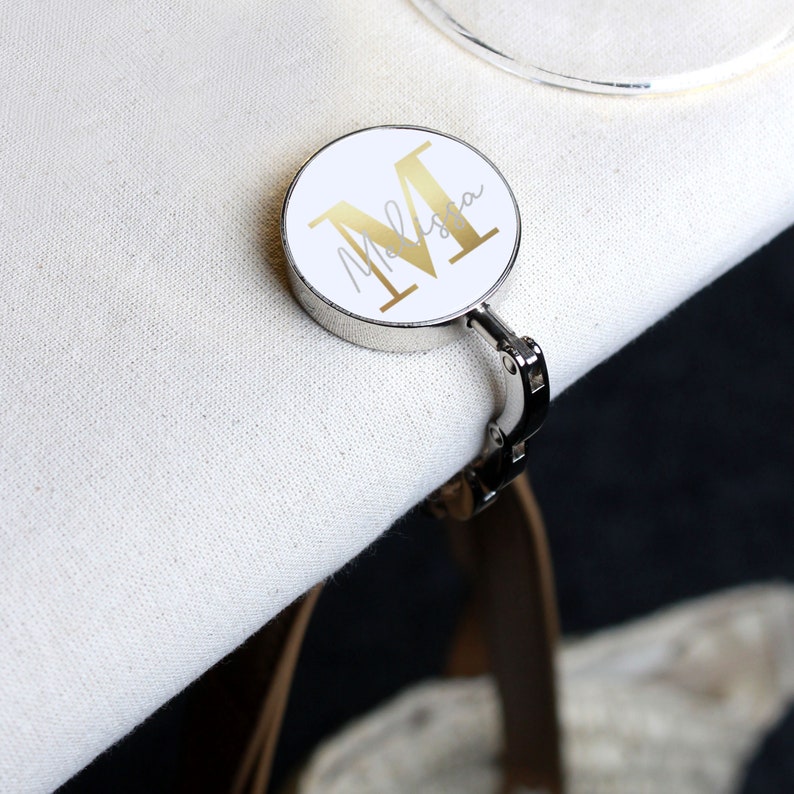 May include: A silver and white personalized bag hook with a gold letter "M" and the name "Melissa" written in cursive.