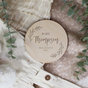 May include: A wooden baby announcement disc with a leafy border. The text reads "Baby Thompson Due August 2025".