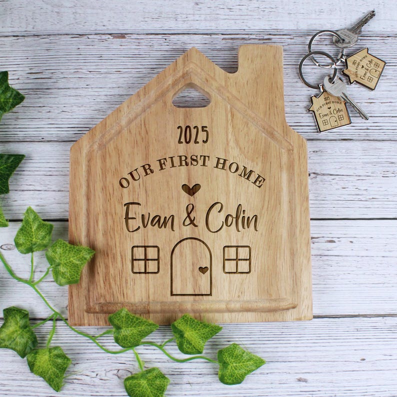 Op de afbeelding: Een houten snijplank in de vorm van een huis met de tekst "2025 Our First Home Evan & Colin" erop gegraveerd. De plank heeft een klein hartvormig uitsnijding in het dak en twee ramen aan elke kant.