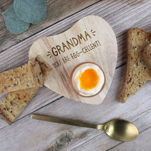 Könnte beinhalten: Ein Eierbecher aus Holz in Herzform mit dem Text "Oma - Du bist egg-cellent!"  Ein weiches Ei befindet sich im Becher, und daneben liegen Toastscheiben.