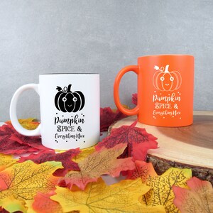 May include: Two ceramic mugs with a fall theme. One is white with a black interior and a black pumpkin graphic. The other is orange with a white pumpkin graphic. Both mugs have the text "Pumpkin Spice & Everything Nice".