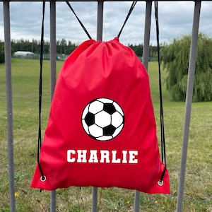 Op de afbeelding: Een rode rugzak met trekkoord met een zwart-witte voetbalafbeelding en de naam "CHARLIE" in witte blokletters. De tas heeft zwarte trekkoorden en metalen oogjes.