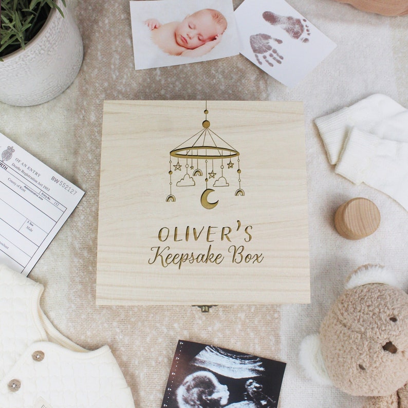 May include: A wooden keepsake box featuring a laser-cut design of a baby mobile with stars, clouds, and a crescent moon. The box is engraved with the text "Oliver's Keepsake Box".