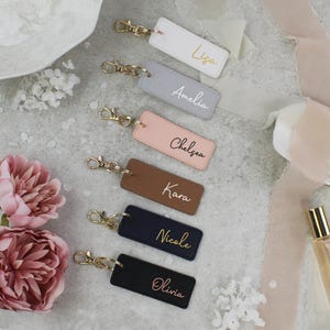 May include: A collection of personalized keychains in various colors, including white, gray, pink, brown, navy, and black. Each keychain features a gold-colored clasp and a name in gold script. The image also includes pink flowers and a perfume bottle.