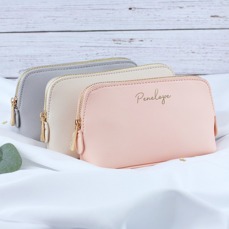 May include: Three zippered pouches in different colors: gray, cream, and pink. The pink pouch has the name "Penelope" written in gold lettering.