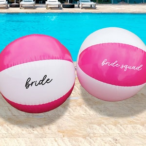 Può includere: Due palloni da spiaggia rosa e bianchi con le parole "bride" e "bride squad" scritte su di essi.