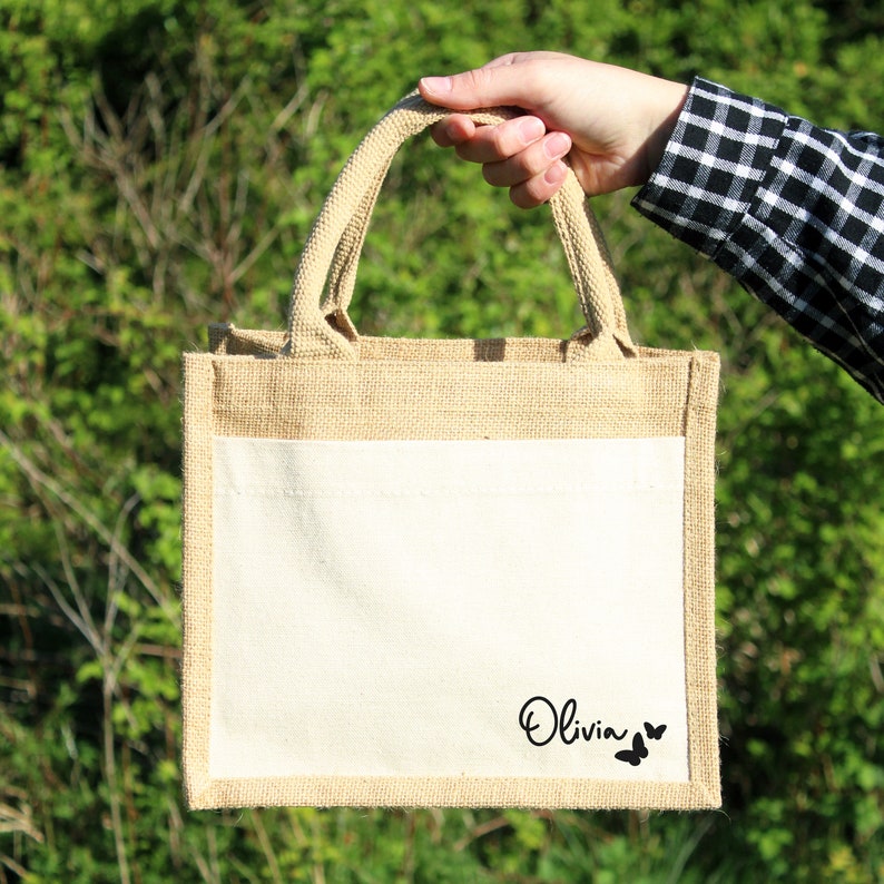 May include: A natural jute tote bag with a white cotton lining. The bag has a single pocket on the front with the name "Olivia" and two butterflies printed in black.