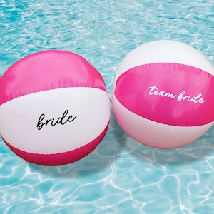 Puede incluir: Dos pelotas de playa rosa y blanca flotando en una piscina. Una pelota dice "bride" y la otra dice "team bride".