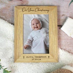 May include: A light-colored wooden picture frame with the text "On Your Christening" at the top. The frame holds a photo of a baby in a white christening gown. The name "OLIVIA THOMPSON" and date "27.03.24" are at the bottom.