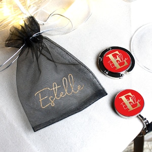 May include: A black mesh bag with the name "Estelle" printed in gold glitter. Two silver and red personalized purse hooks with the letter "E" and the name "Estelle" printed in gold glitter.