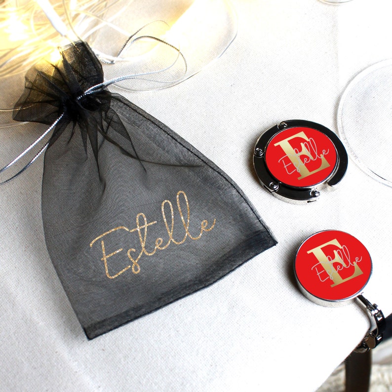 May include: A black mesh bag with the name "Estelle" printed in gold glitter. Two silver and red personalized purse hooks with the letter "E" and the name "Estelle" printed in gold glitter.