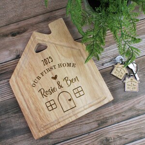 Op de afbeelding: Een houten snijplank in de vorm van een huis met de tekst "2025 OUR FIRST HOME Rosie & Ben" erop gegraveerd. De plank heeft een klein huis-uitsparing met een hart in de deur en ramen.