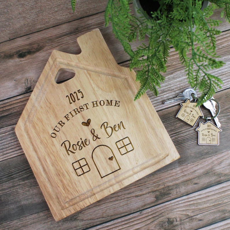 Op de afbeelding: Een houten snijplank in de vorm van een huis met de tekst "2025 OUR FIRST HOME Rosie & Ben" erop gegraveerd. De plank heeft een klein huis-uitsparing met een hart in de deur en ramen.