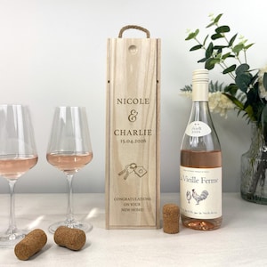 May include: A wooden wine box engraved with "Nicole & Charlie 15.04.2026" and a key and house graphic. Two wine glasses filled with rosé wine and a bottle of rosé wine with a rooster label are next to the box. Two corks are in front of the glasses.