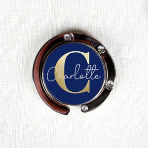 May include: A silver-toned handbag hook with a navy blue circle featuring a gold letter "C" and the name "Charlotte" in white script.