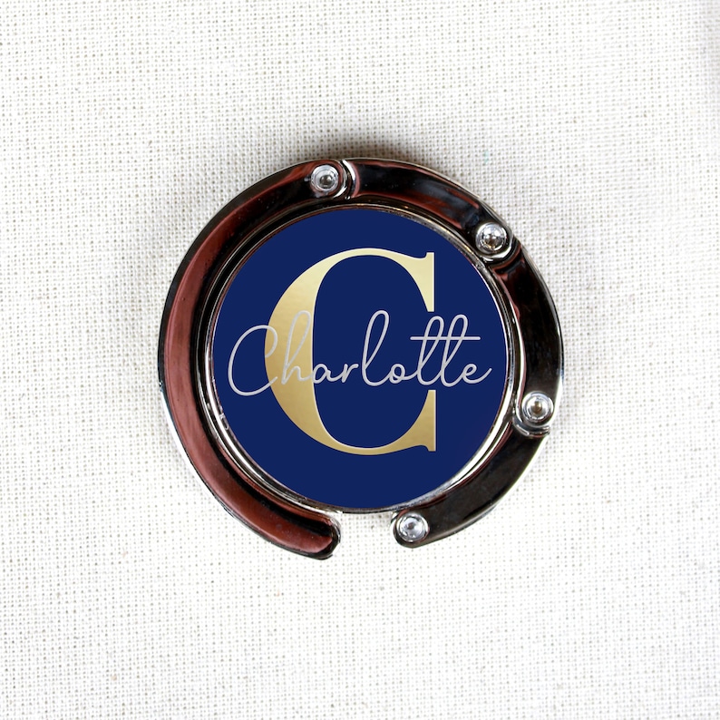 May include: A silver-toned handbag hook with a navy blue circle featuring a gold letter "C" and the name "Charlotte" in white script.