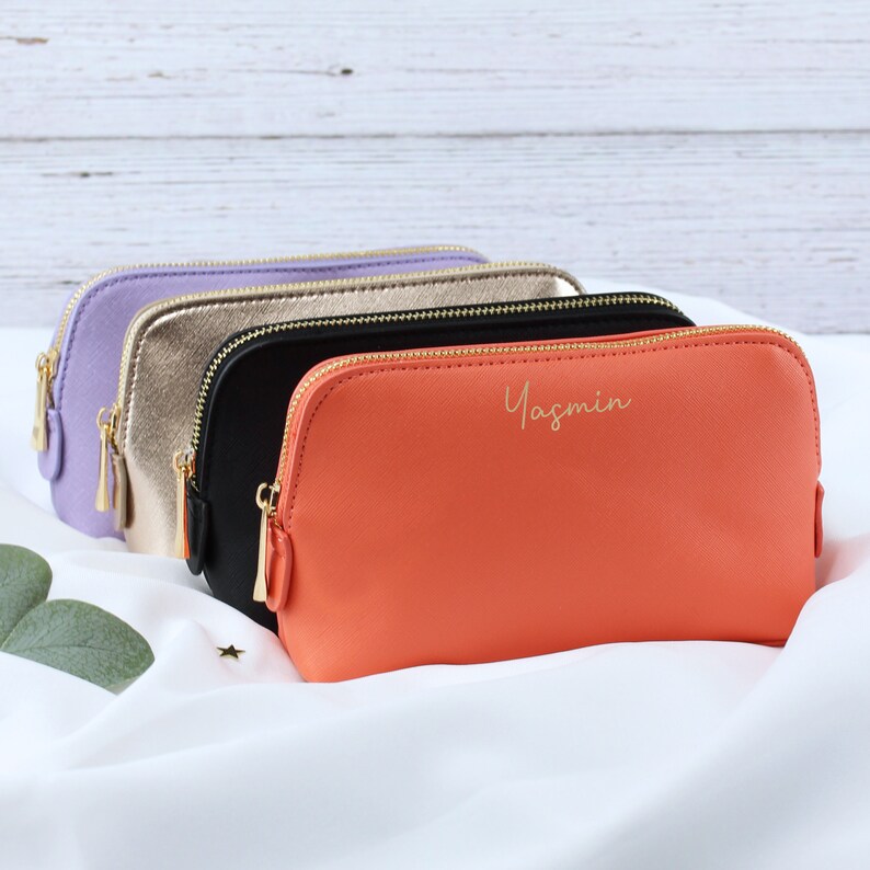May include: Four zippered pouches in different colors: lavender, gold, black, and orange. The orange pouch has the name "Yasmin" written in gold lettering.