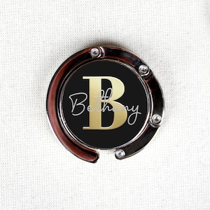May include: A silver-toned bag charm with a black circle featuring a gold letter "B" and the name "Bethany" in a cursive font.