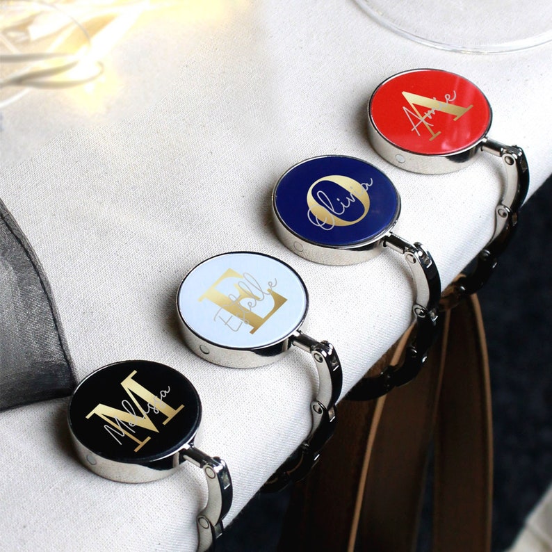 May include: Four personalized table clips with gold lettering on different colored backgrounds. The clips are silver and have a spring-loaded mechanism. The names on the clips are "Olivia", "Belle", "Amelia", and "Melissa".