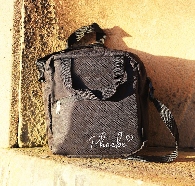 K&ouml;nnte beinhalten: Schwarze Umh&auml;ngetasche mit Rei&szlig;verschlussfach und einer wei&szlig;en Aufschrift "Phoebe" mit einem Herzsymbol.